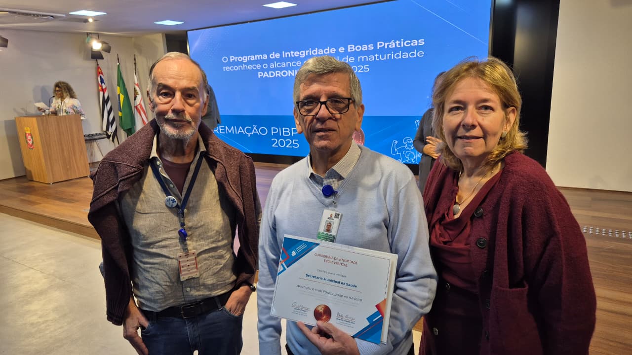 Fotografia registrada em um auditório durante cerimônia de premiação. Em primeiro plano, três pessoas posam para a foto diante de um telão azul. Ao centro, um homem de cabelos grisalhos usa suéter claro e segura um certificado de reconhecimento da Secretaria Municipal da Saúde. À esquerda, um homem de barba grisalha veste camisa clara e casaco marrom apoiado sobre os ombros. À direita, uma mulher de cabelos loiros curtos usa conjunto em tom vinho. Ao fundo, há um palco com púlpito, bandeiras do Brasil, do Estado de São Paulo e do Município de São Paulo, além de uma pessoa ao microfone. No telão, aparece a identidade visual da premiação do Programa de Integridade e Boas Práticas (PIBP) 2025. 