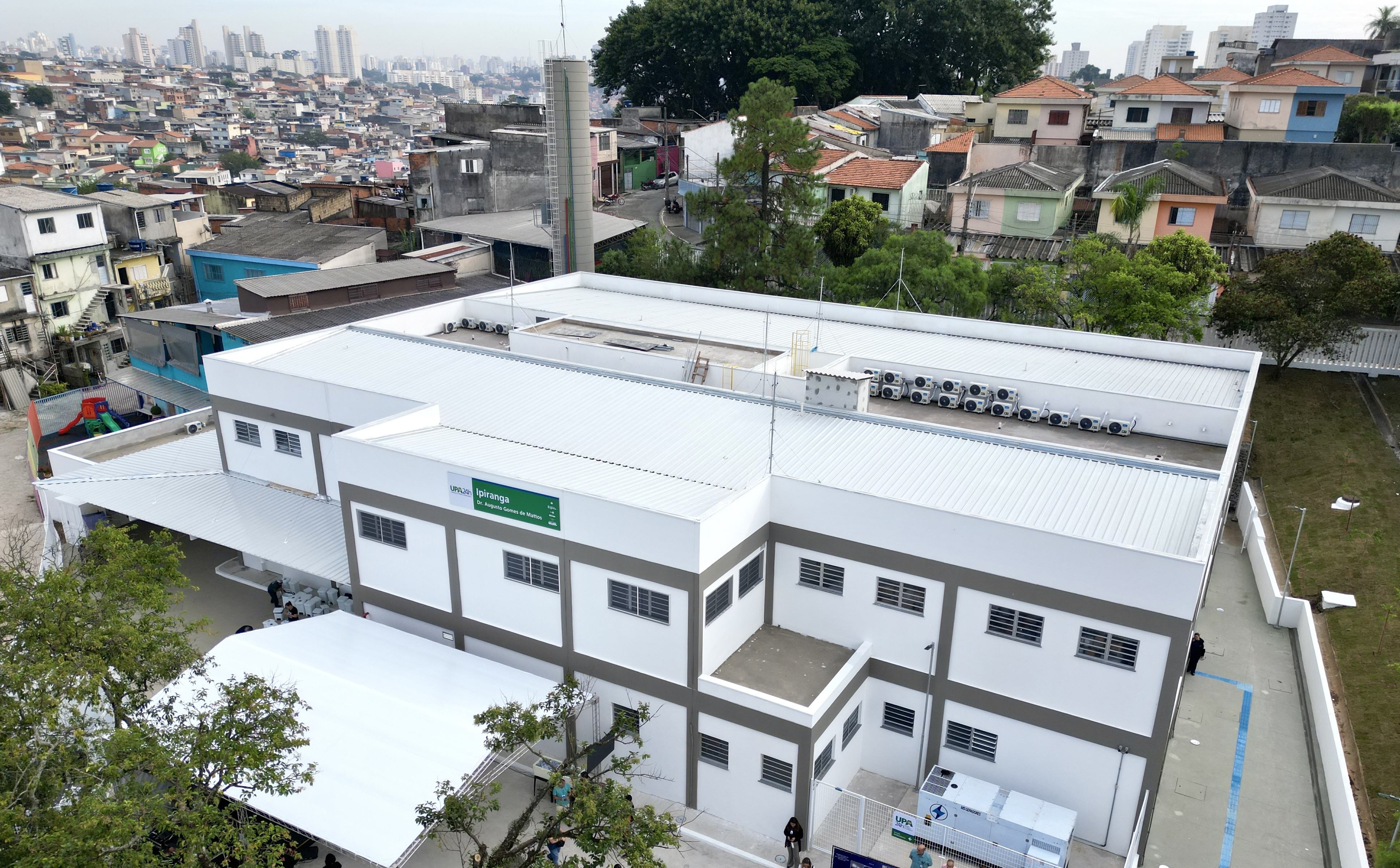  A imagem mostra uma vista aérea da Unidade de Pronto Atendimento (UPA) Ipiranga, que é um prédio grande, moderno e de cor clara. A estrutura tem formato retangular, com telhado metálico branco e várias janelas distribuídas pelas paredes externas. Na lateral do edifício, há uma cobertura branca onde algumas pessoas estão reunidas, possivelmente aguardando atendimento. Próximo a essa área, é possível ver uma pequena área com brinquedos coloridos, sugerindo um espaço infantil. No topo do prédio, há diversos equipamentos instalados, como aparelhos de ar-condicionado alinhados e uma estrutura vertical que parece ser um reservatório ou torre técnica. Ao redor do edifício, há árvores e áreas abertas. O entorno mostra um bairro bastante denso, com muitas casas pequenas e construções próximas umas das outras, além de prédios altos ao fundo, indicando uma área urbana extensa.