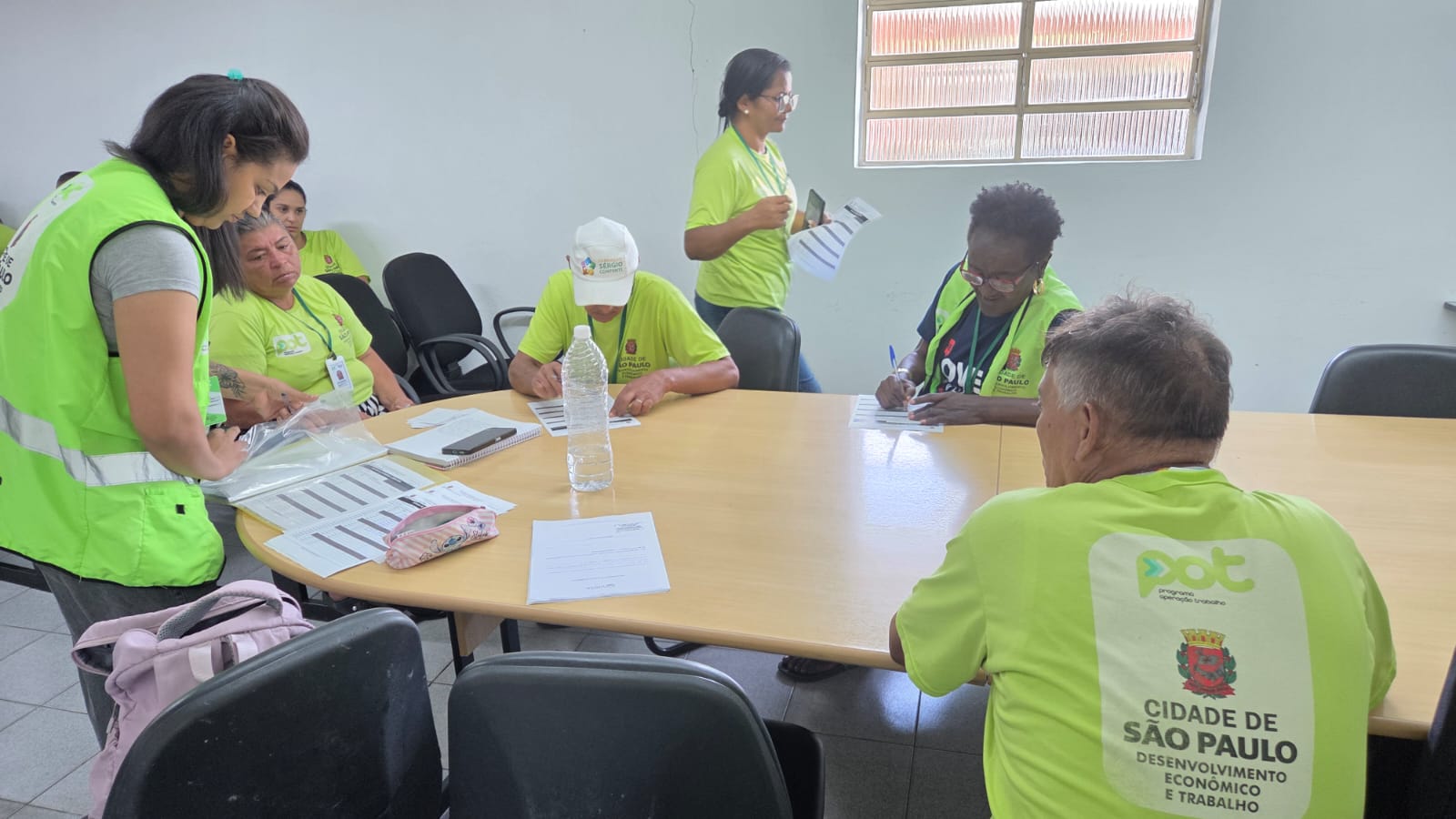 A imagem mostra um grupo de pessoas reunidas em uma sala simples, sentadas ao redor de uma mesa oval de madeira clara. A maioria veste camisetas verdes com identificação do POT. À esquerda, uma mulher em pé, também de verde, está inclinada sobre a mesa, organizando ou entregando materiais dentro de um saco plástico. Ao redor da mesa, outras pessoas — homens e mulheres, em sua maioria adultos e idosos — estão sentadas, concentradas em preencher formulários com caneta.