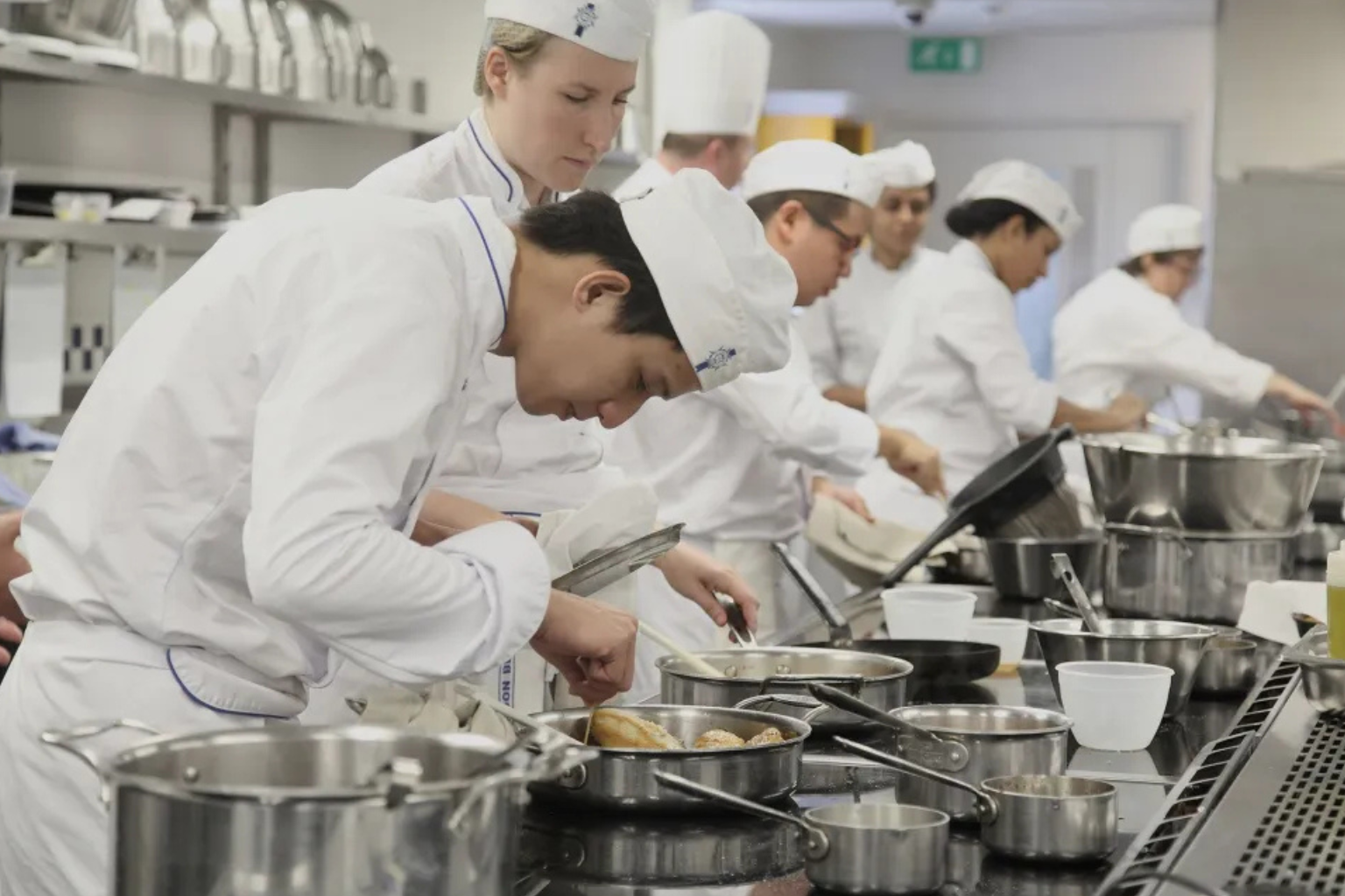 Alunos de culinária com aventais brancos cozinhando.