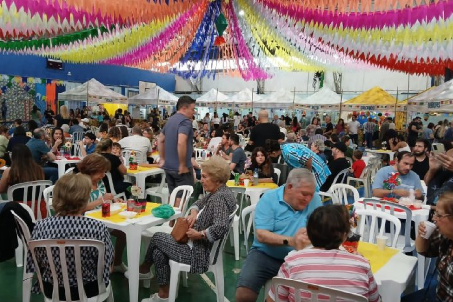  Pessoas reunidas na festa junina repleta de bandeiras coloridas na decoração.