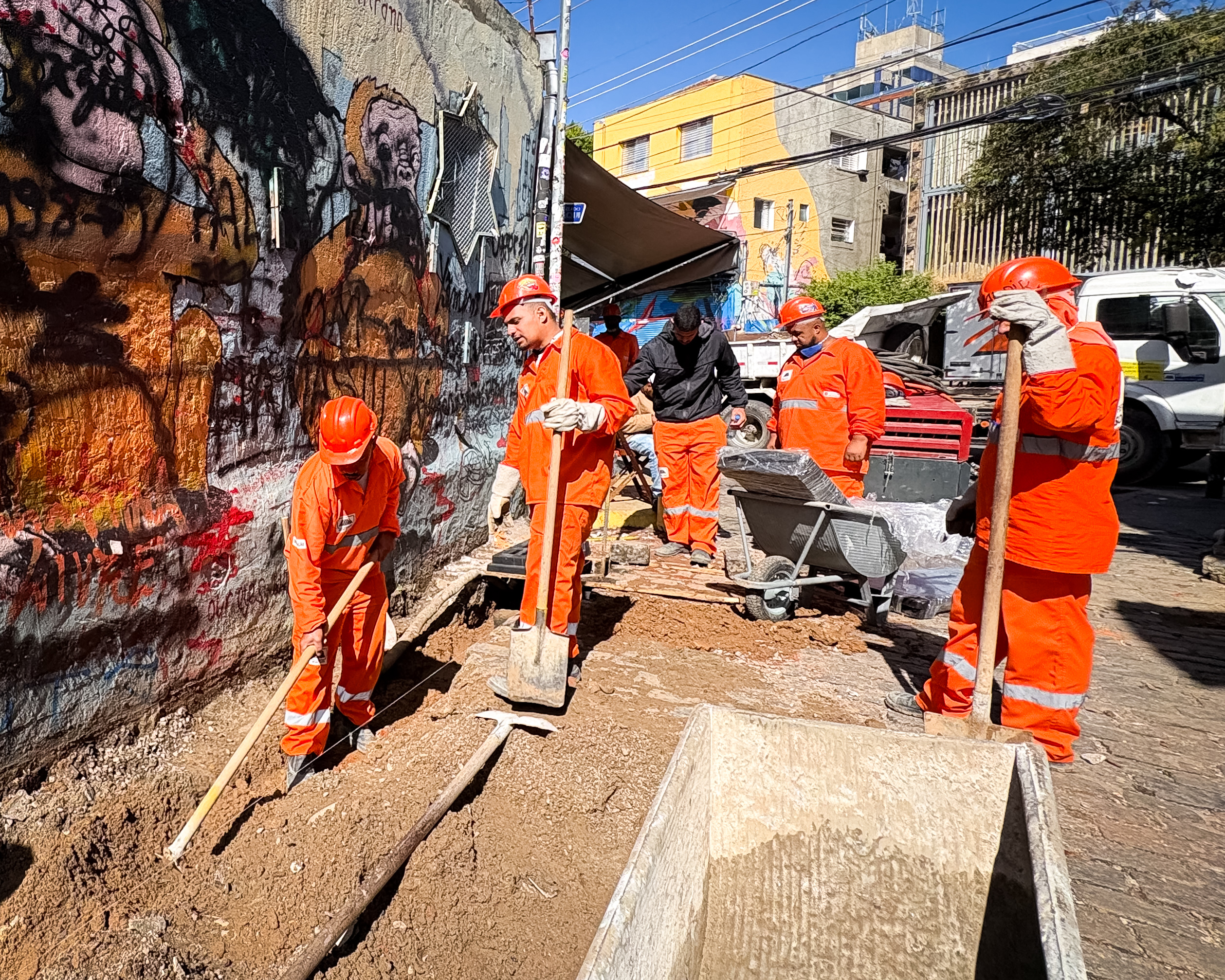 Imagem mostra equipes realizando obras de requalificação no Beco do Batman, em São Paulo. Trabalhadores uniformizados em laranja utilizam ferramentas e carrinhos de obra ao lado dos murais de grafite característicos do local. A via está parcialmente escavada para execução das melhorias de infraestrutura e acessibilidade. Ao fundo, aparecem edifícios, postes e a paisagem urbana da região da Vila Madalena sob céu azul.