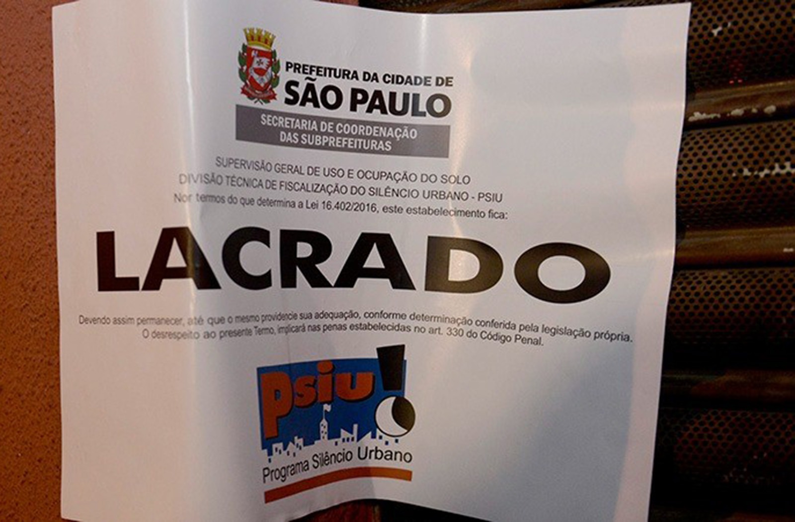Imagem mostra um aviso de lacração emitido pela Prefeitura da Cidade de São Paulo, fixado em um estabelecimento. O documento pertence ao Programa Silêncio Urbano (PSIU) e informa a interdição do local por descumprimento da legislação relacionada ao controle de ruído urbano. Em destaque, aparece a palavra “LACRADO” em letras grandes no centro do aviso.